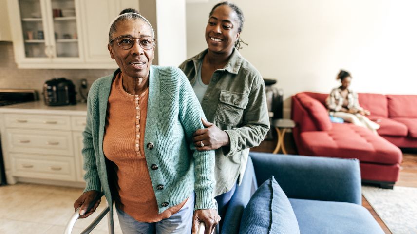 An older woman with glasses, is walking in her home using a walker, smiling, with a younger woman standing behind her, smiling offering support with a gentle hand on her arm. In the background a young girl is sitting on a red couch in the living room.
