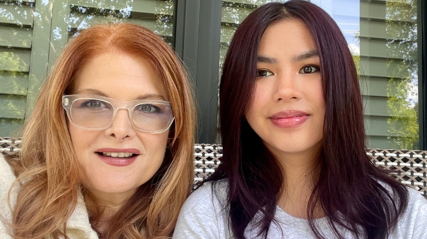 Selfie of Maria and her daughter smiling, sitting outdoors, a window reflecting tree foliage is behind them in the background.