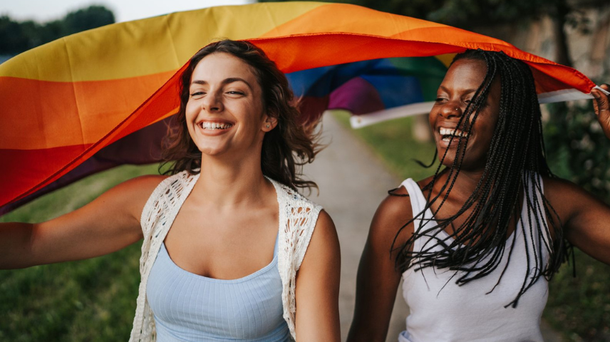 Happy LGBTQ+ Pride Month - celebrating diversity and advocating for Health Equity. Two smiling women running under a colorful rainbow flag. The woman on the left has dark hair and wears a light blue tank top and white knit vest, and the woman on the right has long flowing black dreadlocks and wears a white tank top. They are outdoors with greenery in the background.